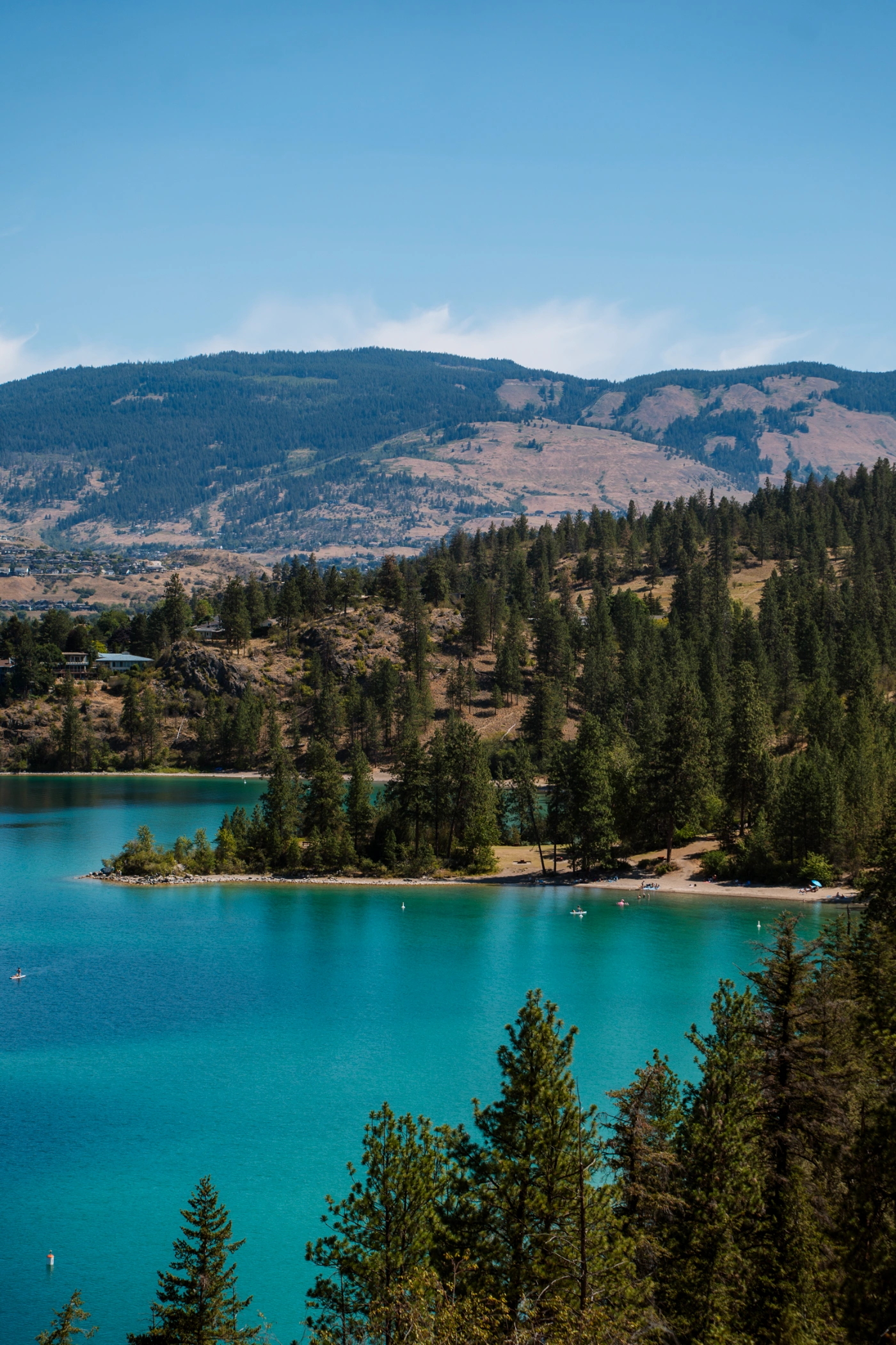 A woman sits on a large boulder at the top of a mountain looking down at the massive stretch of Okanagan Lake