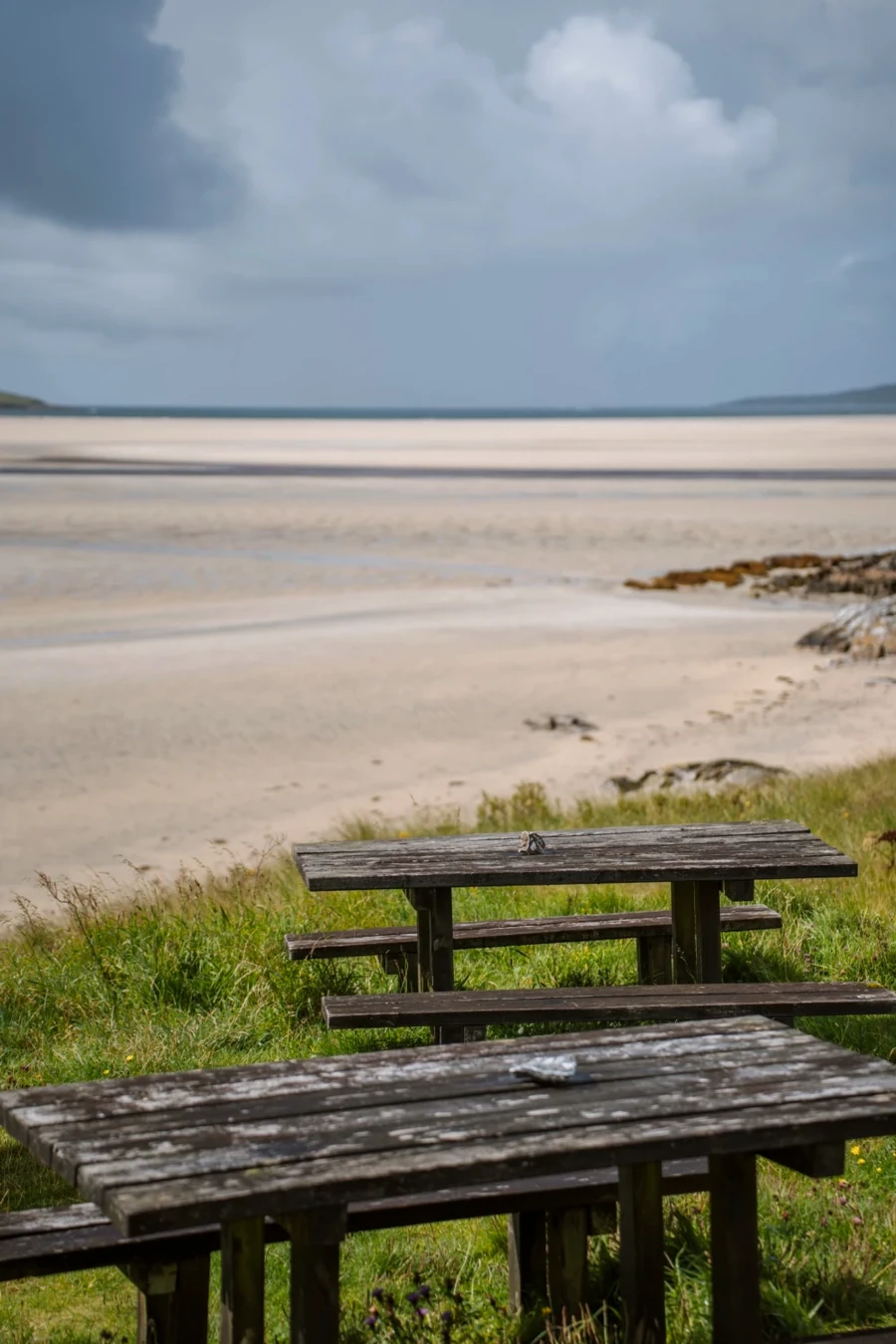 Two weathered picnic tables on a grassy patch above a large white sand beach