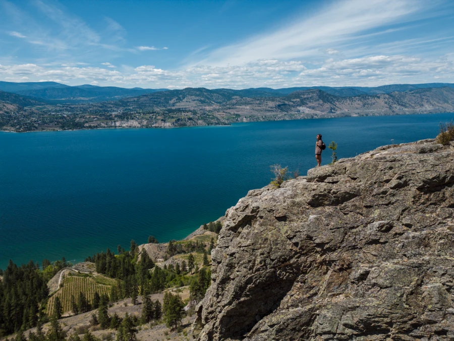 A woman stands on the edge of a massive rocky cliff at the end of the Lady Bug Trail near Little Tunnel with Okanagan Lake stretching as far as the eye can see below.