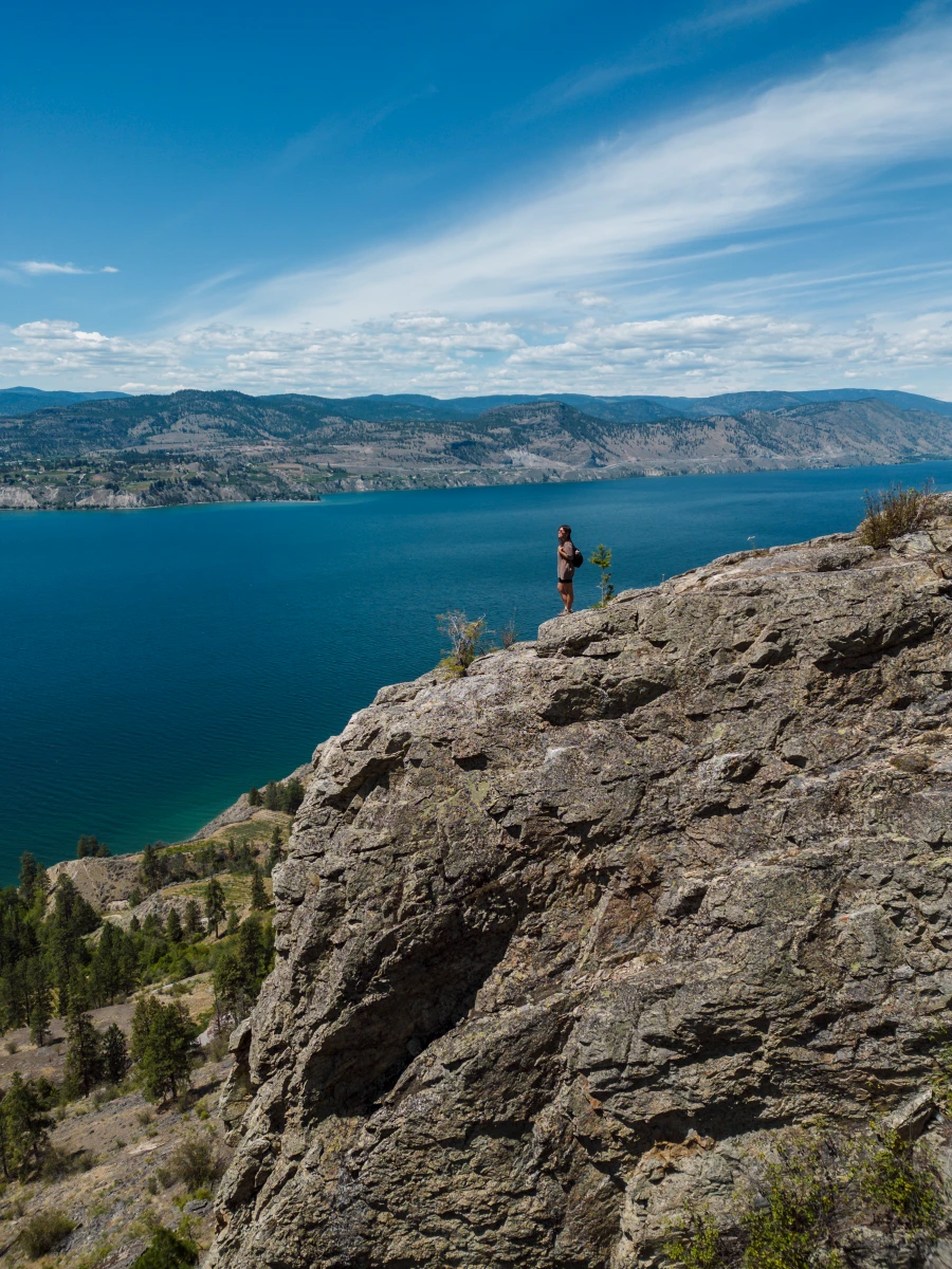 A woman stands on the edge of a massive rocky cliff at the end of the Lady Bug Trail near Little Tunnel with Okanagan Lake stretching as far as the eye can see below.