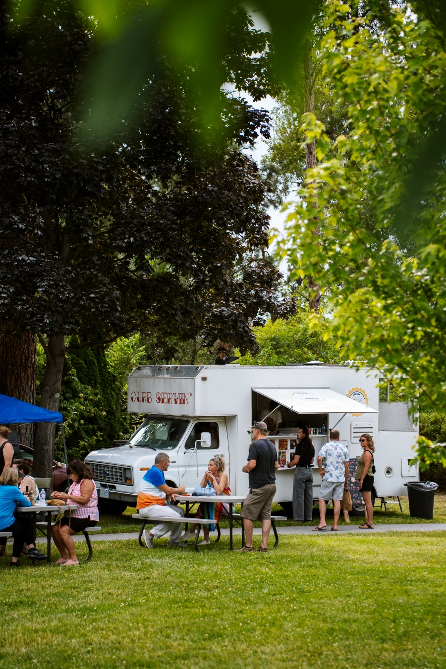 Several picnic tables are set up on a grassy area next to a paved pathway that leads past several food trucks at the Naramata Farmers’ Market.