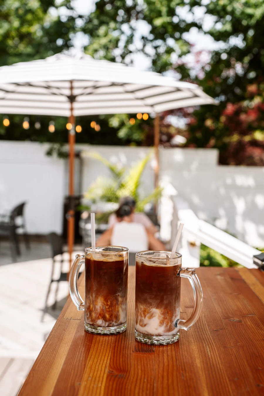 Two iced coffees in glass mugs with straws sit on a polished wood table on a patio with several tables and umbrellas behind it.