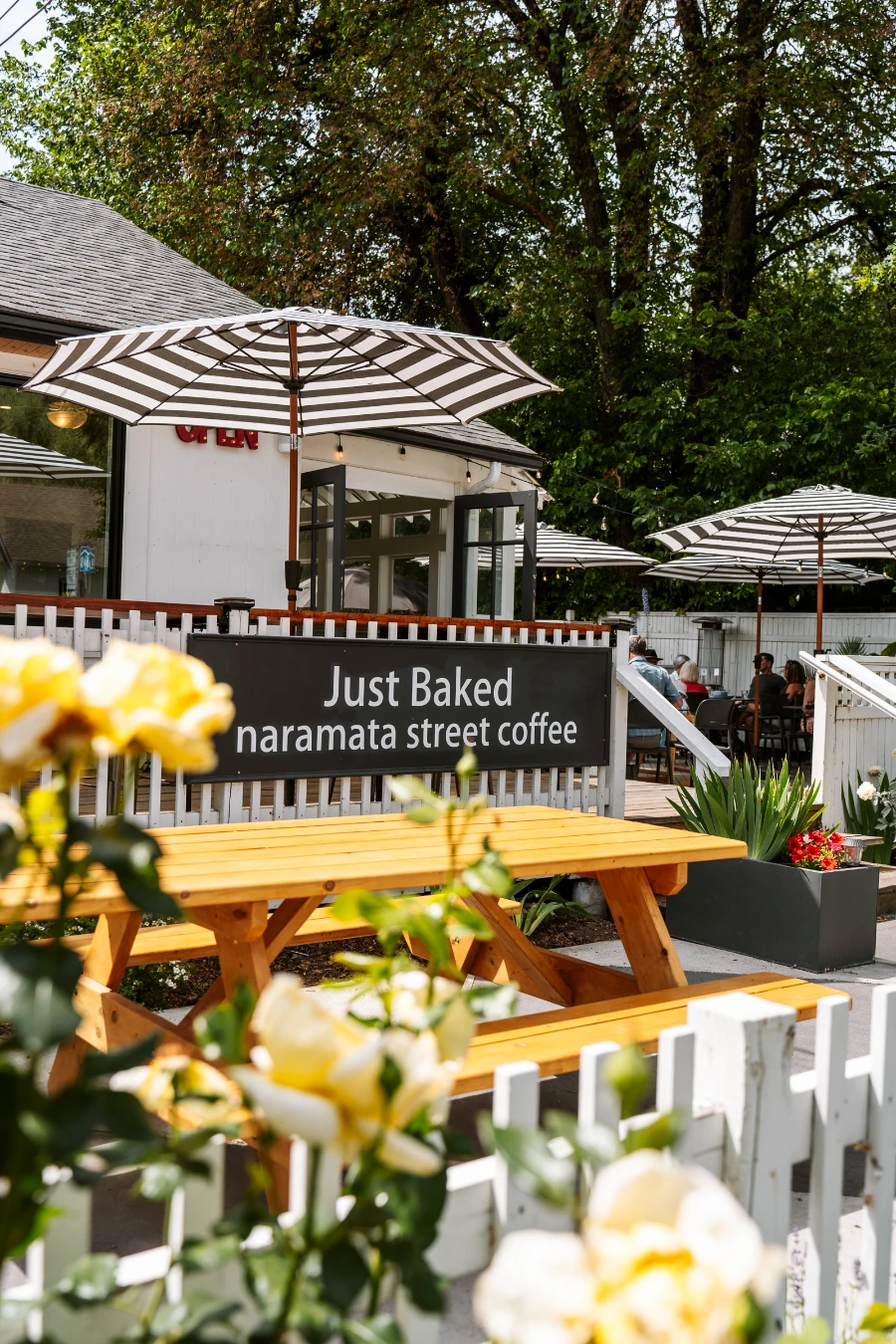 Blooming flowers frame the foreground, with a large patio with several picnic tables and round tables with umbrellas making up the midground, with a fence running through with a sign hung on it reading ‘Just Baked Naramata Street Coffee’
