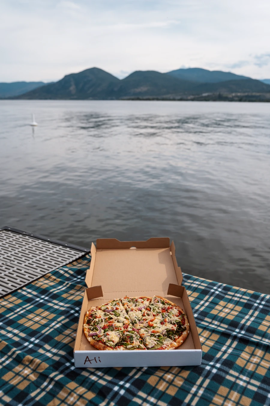 A full pizza inside an open cardboard box lying on a picnic blanket set up on a dock that is suspended above the water of Okanagan Lake.