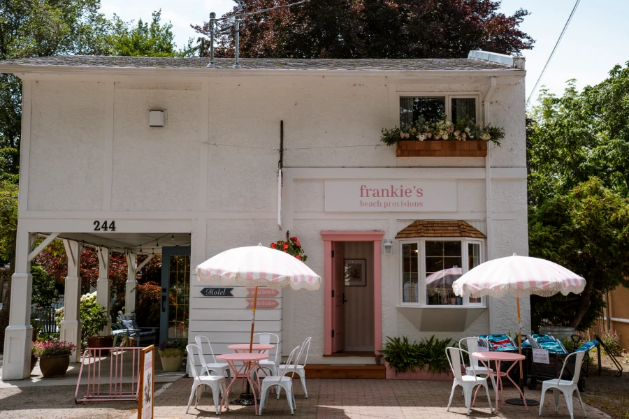 A quaint white building with a few small tables set up in front of it with a sign that reads in pink letters ‘Frankie’s Beach Provisions’