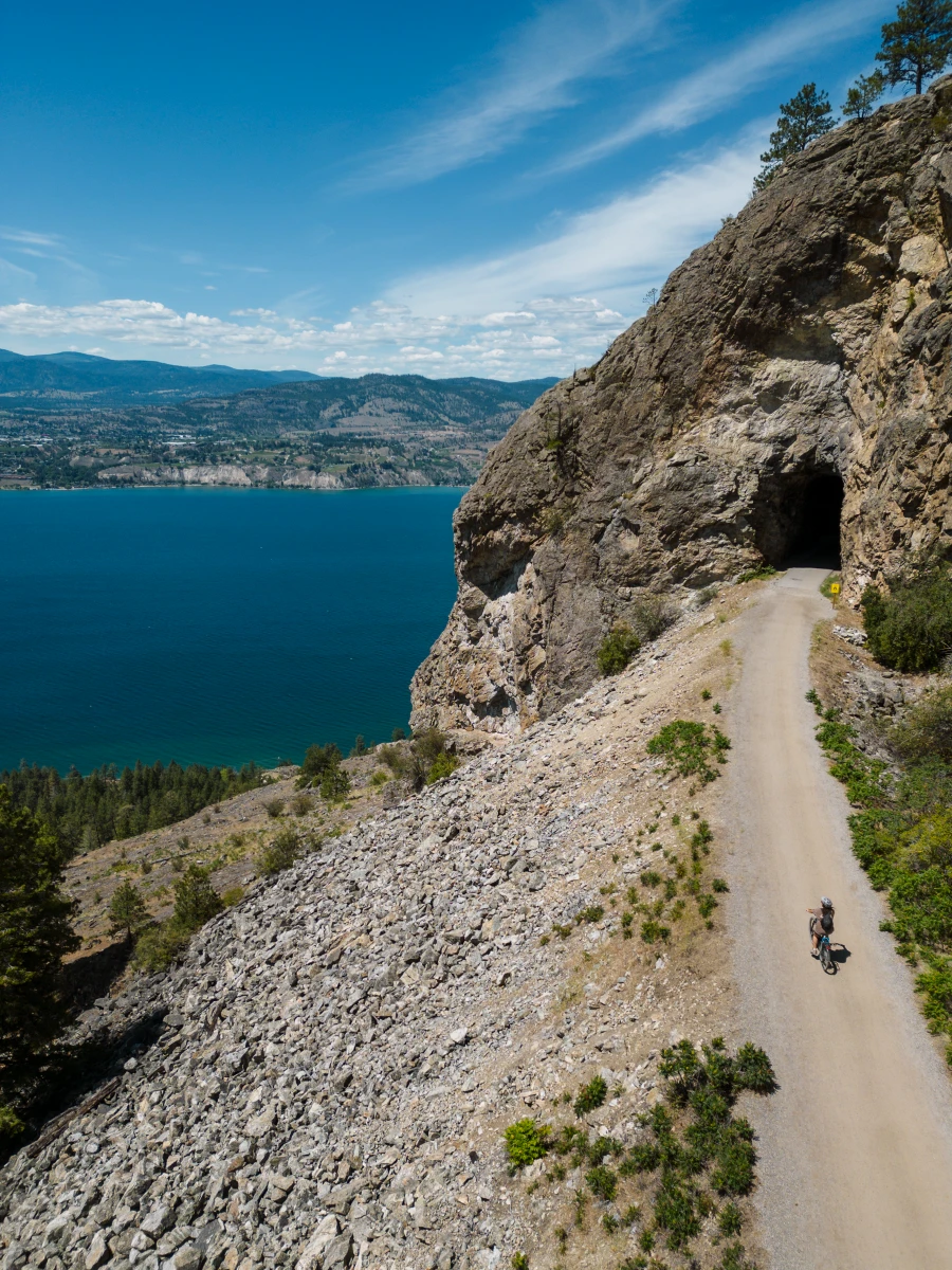 A woman riding an E-bike along the gravel KVR trail heading into a dark tunnel known as ‘Little Tunnel’ with the striking blue of Okanagan Lake far below the path to the left.