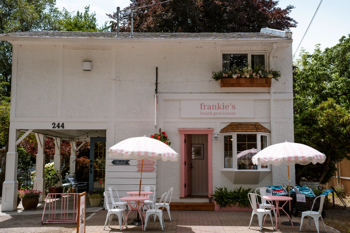 A quaint white building with a few small tables set up in front of it with a sign that reads in pink letters ‘Frankie’s Beach Provisions’