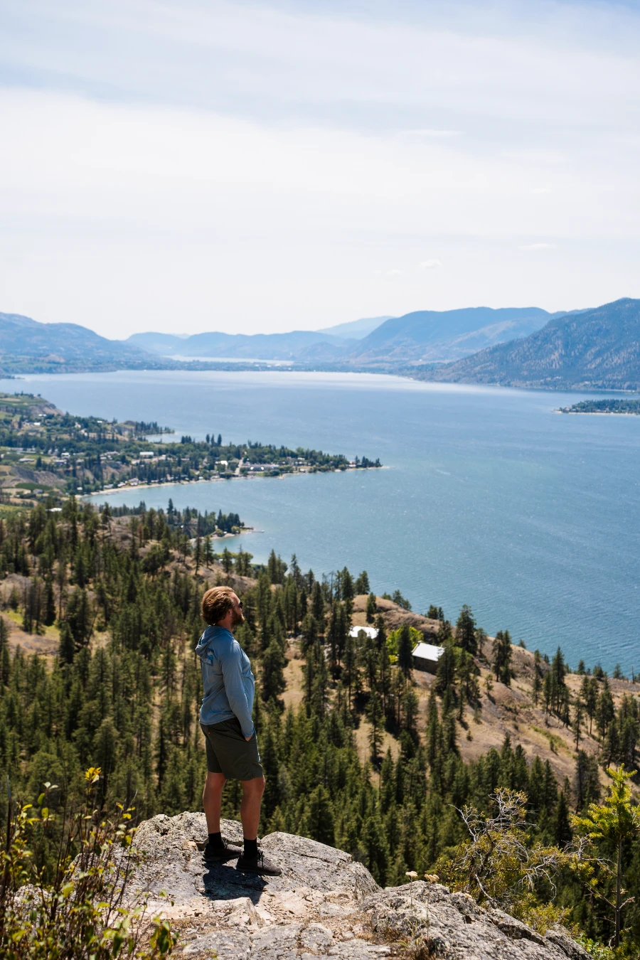 A man stands on the edge of a rocky cliff at the end of the Lady Bug Trail near Little Tunnel, looking down at the Naramata Bench and Okanagan Lake.