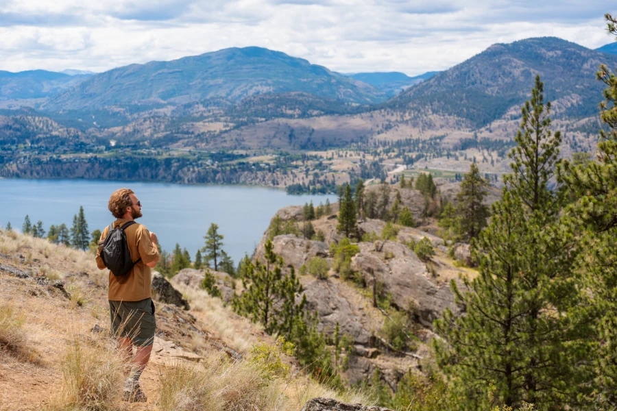 A man stands on a hiking trail surrounded by rocky bluffs and dry yellow tufts of grass with Okanagan Lake and distant mountains visible in the background.