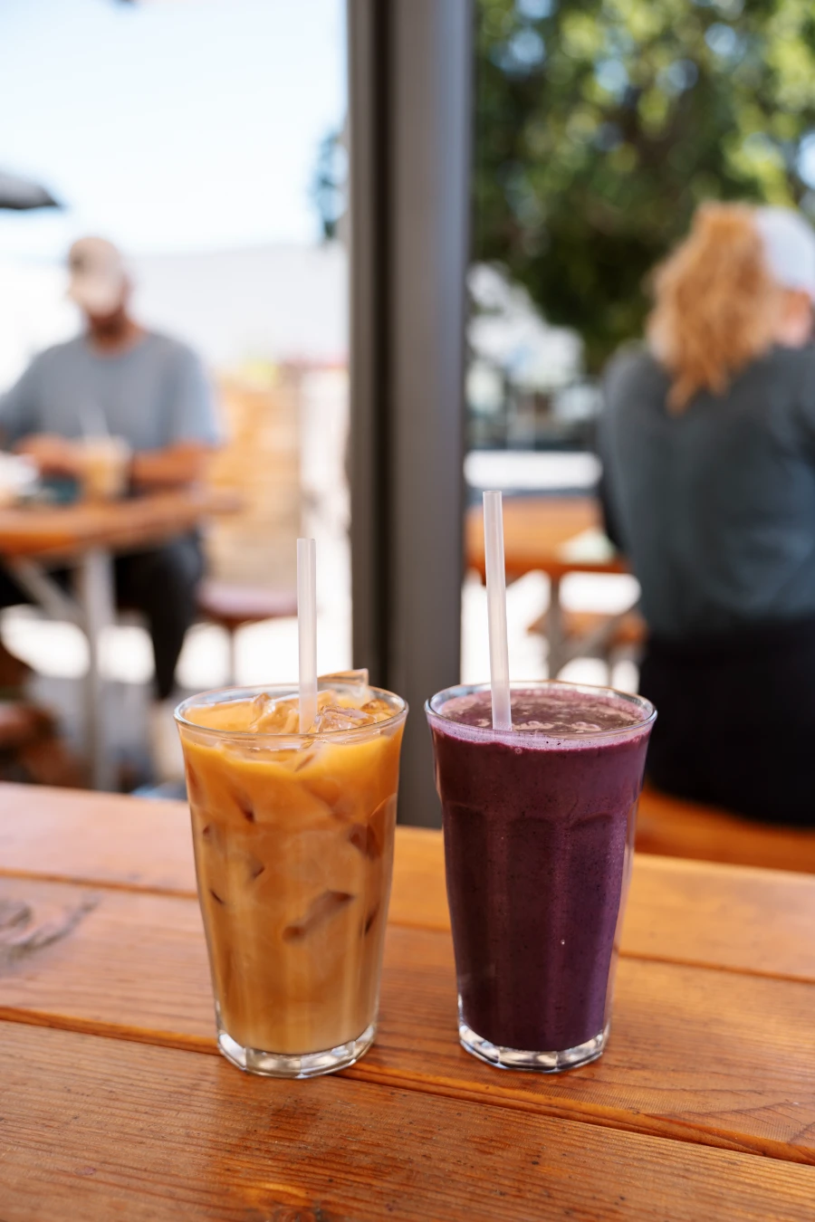 Two tall glasses sit on a wooden picnic table. One is filled with iced coffee and the other with a purple fruit smoothie.
