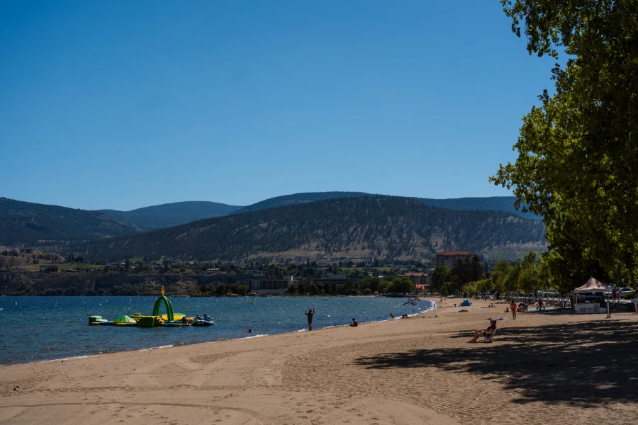The sandy shores of Okanagan Lake Beach with inflatable playgrounds on the water and layers of mountains on the horizon.