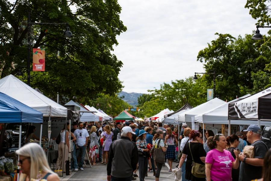 A large crowd fills the street shoulder to shoulder with farmers’ market booths lining either side of the road.