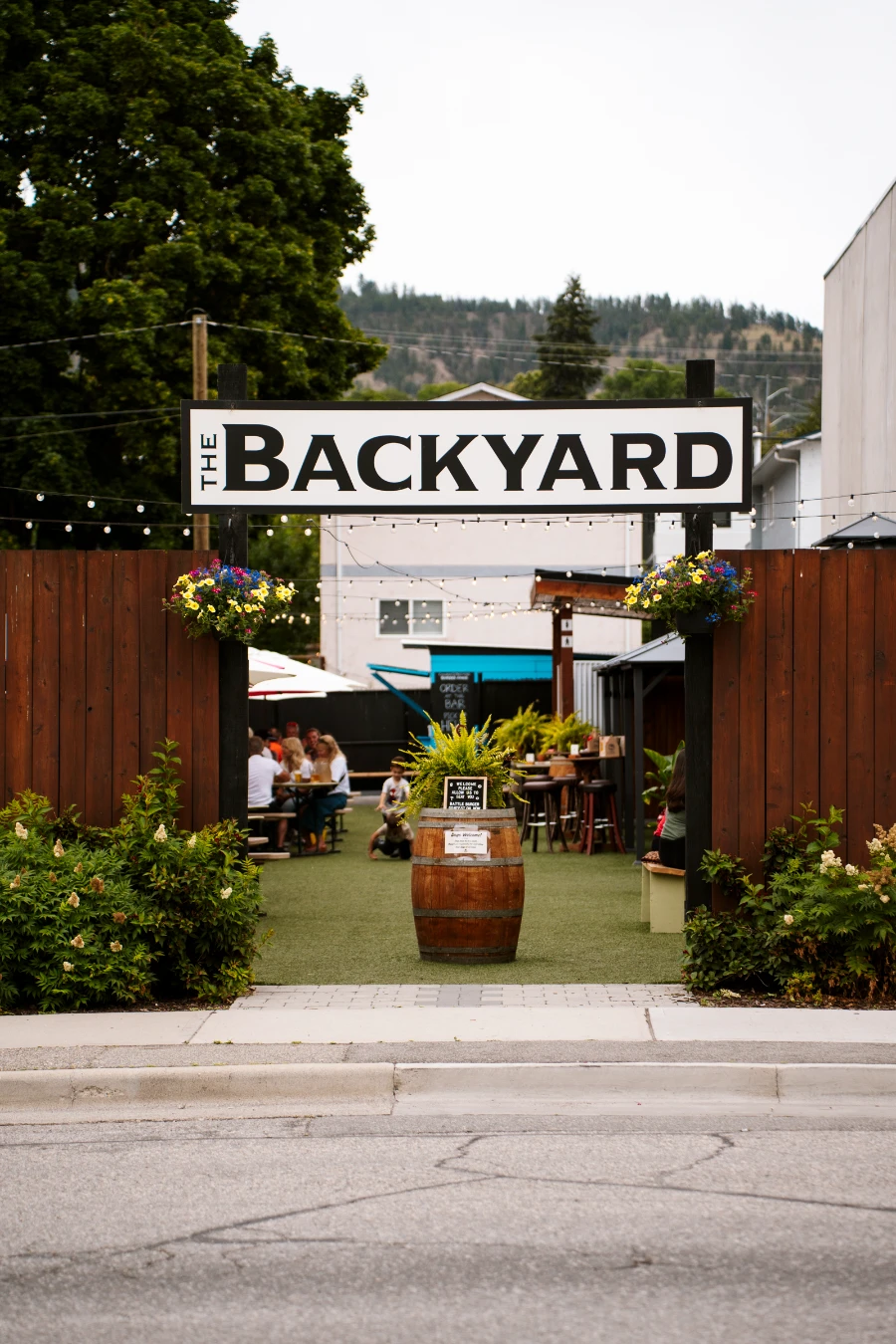 A large sign reads ‘Backyard’ suspended over a gap in a large wooden fence, creating an entryway into a grassy area with tables and umbrellas at Cannery Brewing in Penticton.
