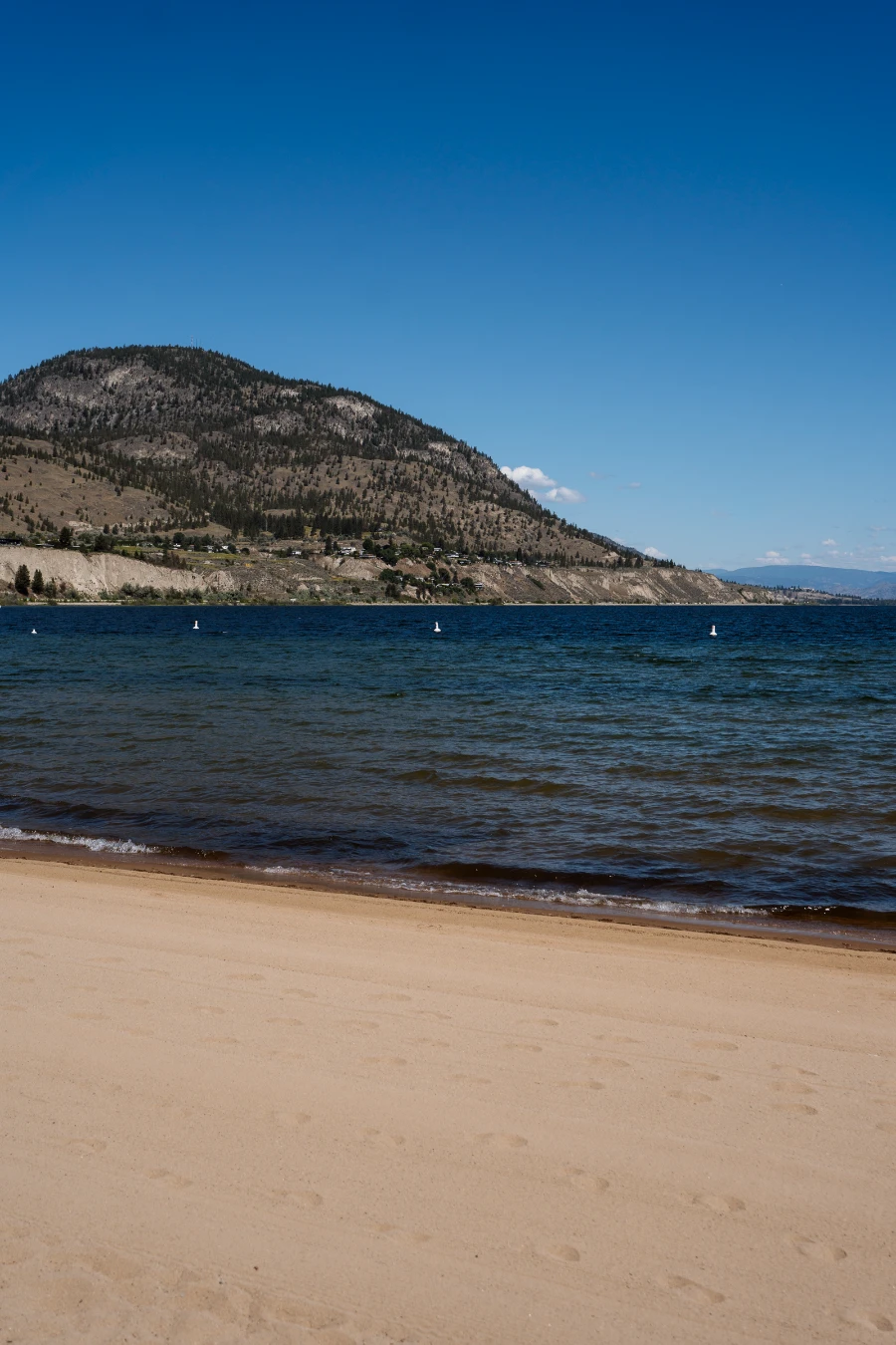 A freshly combed white sand beach with gentle waves lapping the shore from Lake Okanagan with a sparsely forested hill in the distance