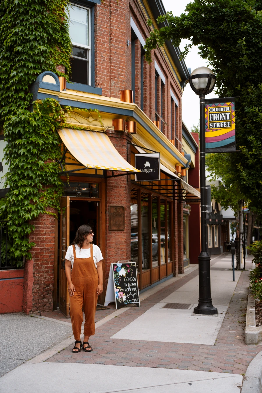 A woman stands on a sidewalk in Penticton in front of an old red-stone building with a colourful awning next to a lamppost that holds a sign that reads ‘Colourful Front Street’.