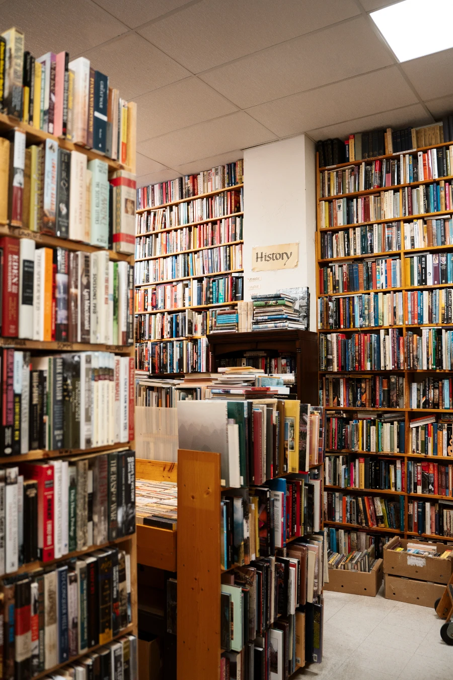 Rows of books and boxes of books on the floor fill a room, and the only blank space left on the wall has a sign written on brown paper that reads ‘History’.