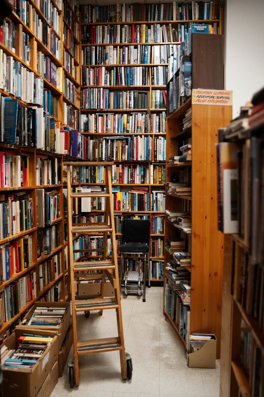 Shelves of books cover the wall and boxes of books are scattered across the floor, and a wooden ladder sits in the middle of the mess.