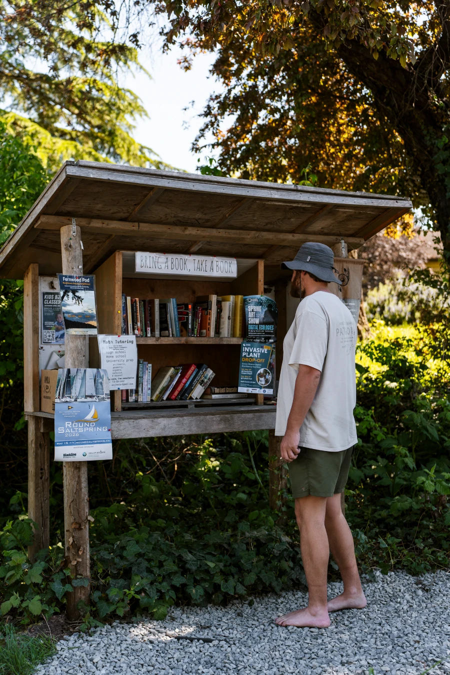 A man with bare feet and a sunhat looks at the collection of books stocked in a small outdoor library