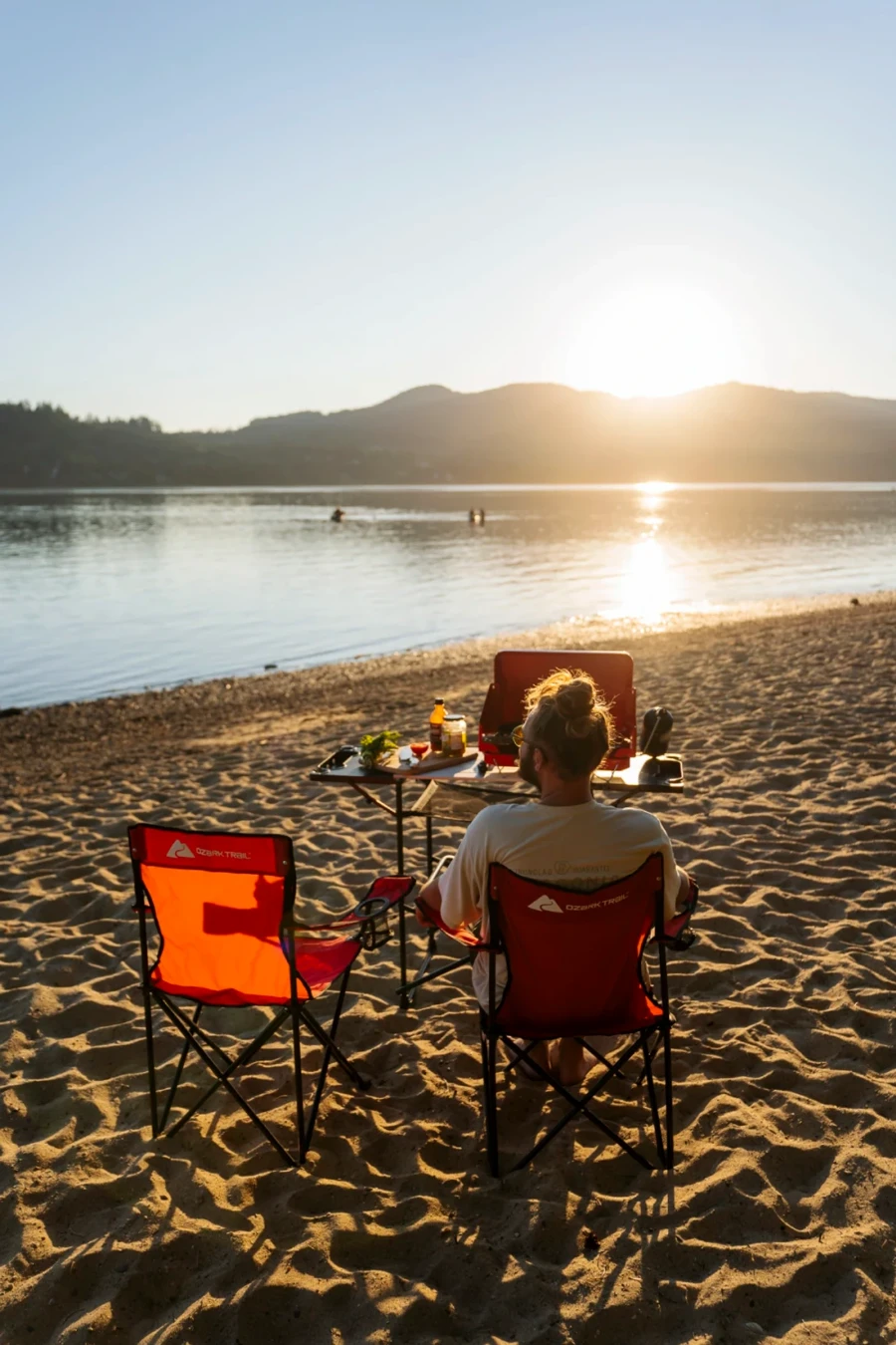 A man sits in a red camp chair in front of a table holding a camp stove on a sandy beach during sunset
