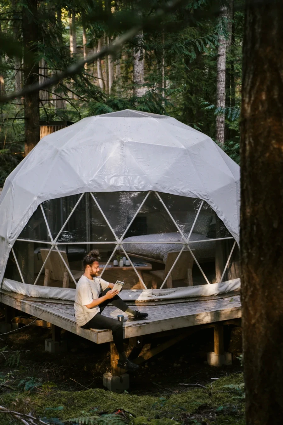 man reading a book while siting on a wooden platform that is shared with a white glamping dome