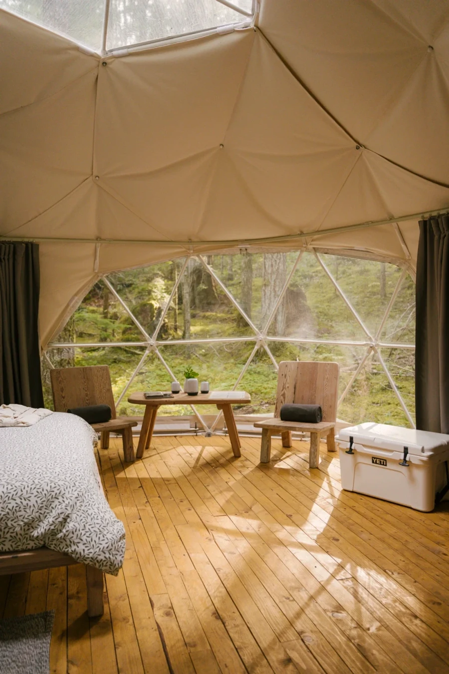 The inside of a white tarp glamping dome showing the bed, table, chairs, and yeti cooler.