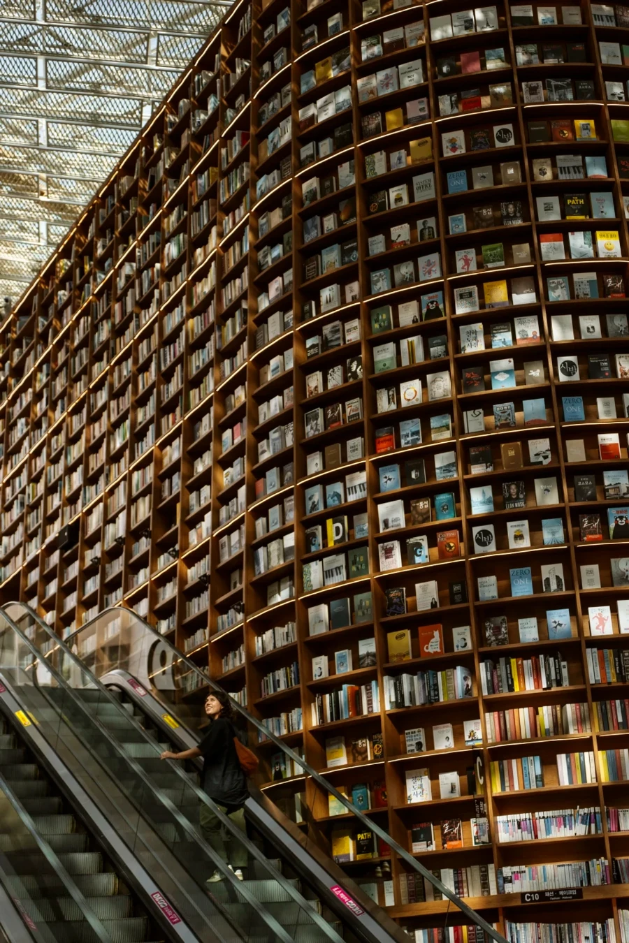 A woman rides an escalator that rises past a massive shelf holding hundreds of books in Starfield Library, Seoul