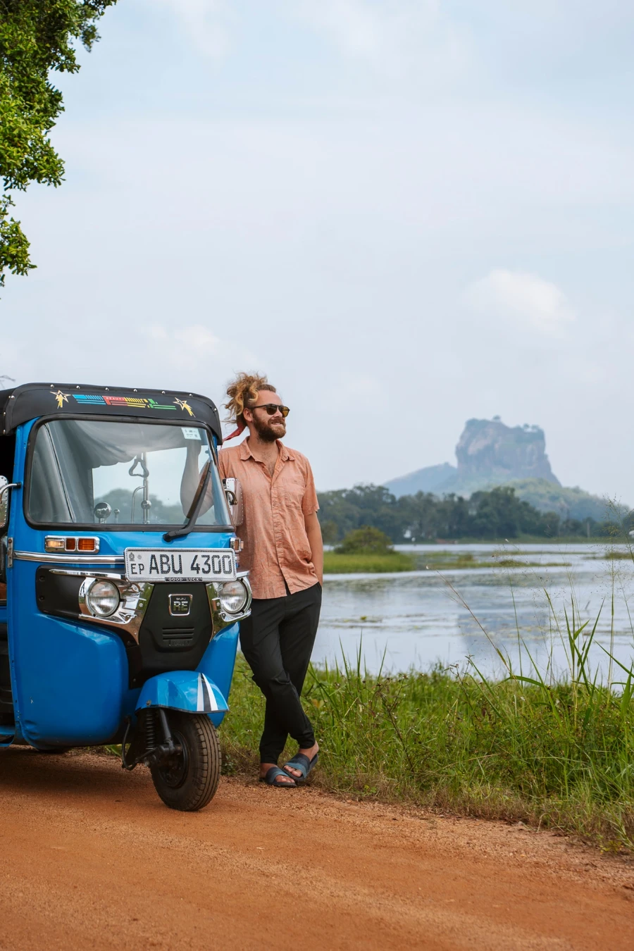 Man stands leaning on a blue tuktuk on a dirt path with a lake and Lions Rock behind him in Sigiriya