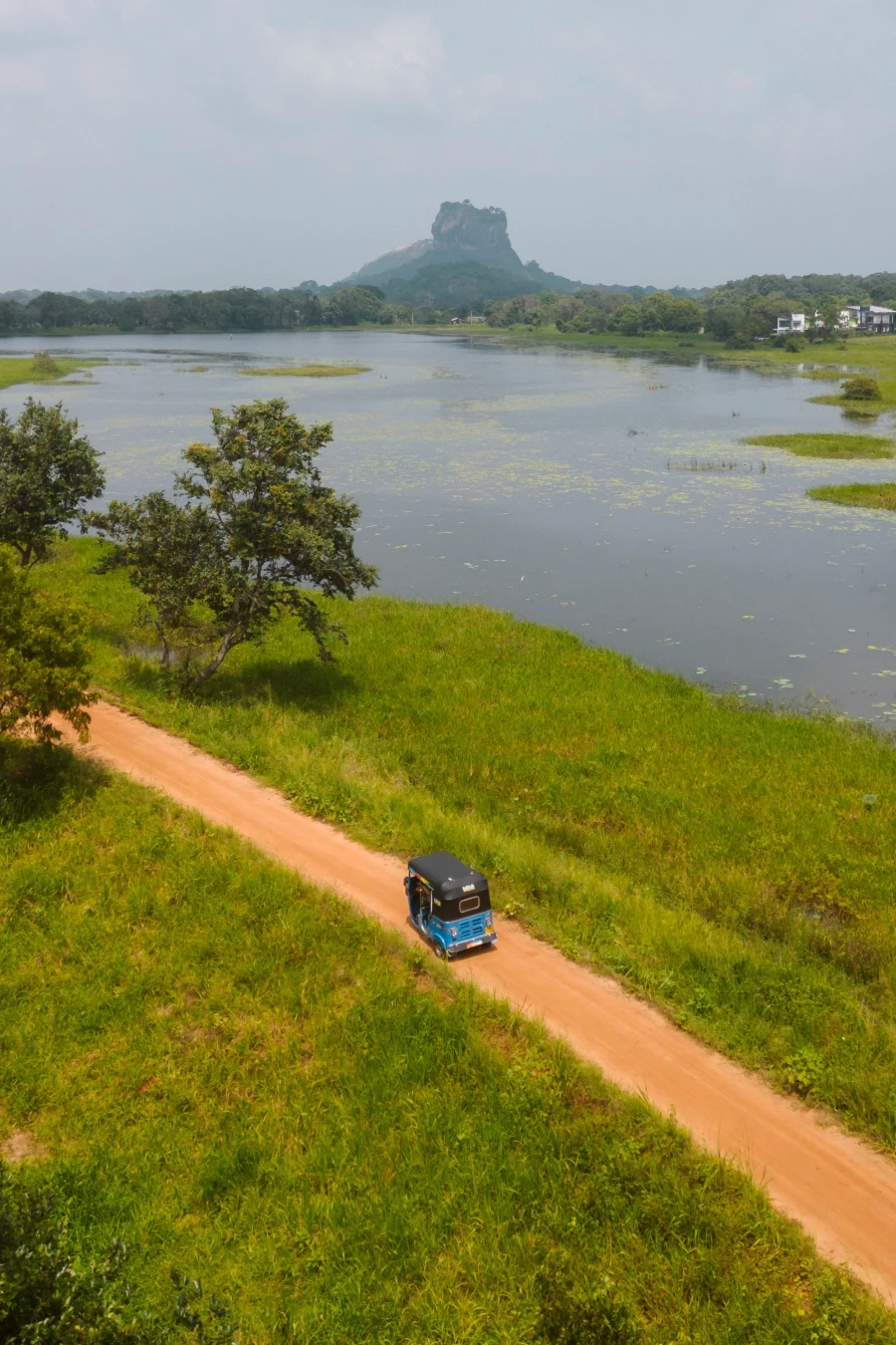 A blue tuktuk drives along a narrow dirt road next to a large lake with Lions Rock visible in the distance