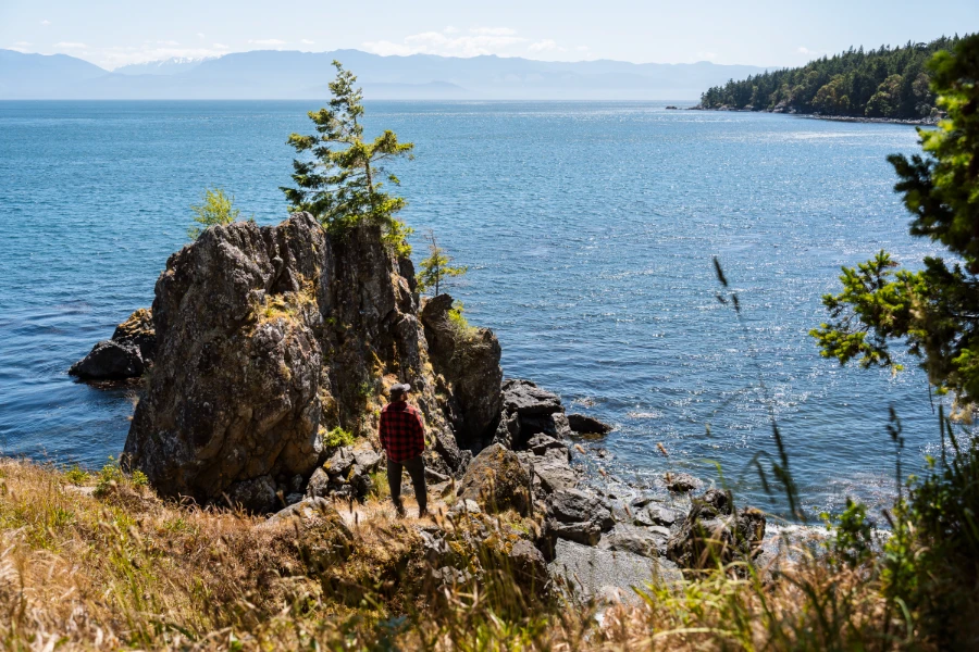 A man stands at the edge of a cliff above a sparkling, calm sea of blue water in East Sooke Regional Park