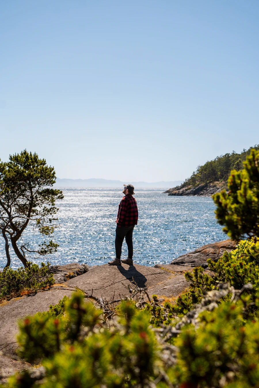 A man stands at the end of a rocky outcrop framed by green trees and standing above a calm sparkling sea in East Sooke Regional Park