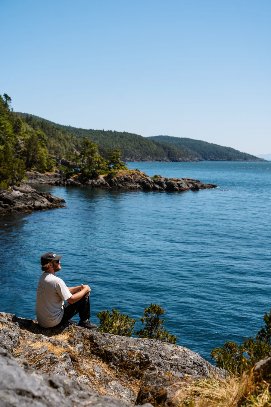 A man sits on a rocky cliff above a calm sea with a rocky and tree-covered coastline stretching out behind him.