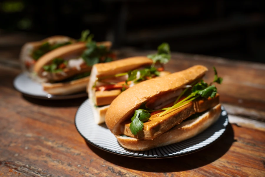 Two Tofu Bahn Mi Sandwiches, cut in half on two small serving plates, sit on a wooden picnic table.