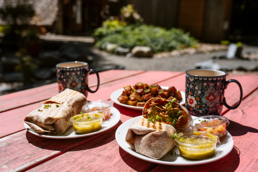 Two plates of burritos and a plate of hashbrowns sitting on a wooden picnic table next to two coffee mugs at Shirley Delicious