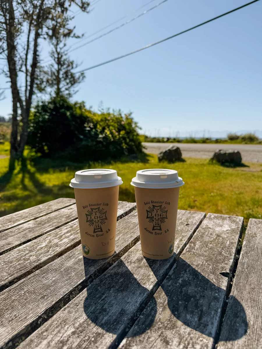 Two takeaway coffee cups on a wooden picnic table with a beach across the highway behind them