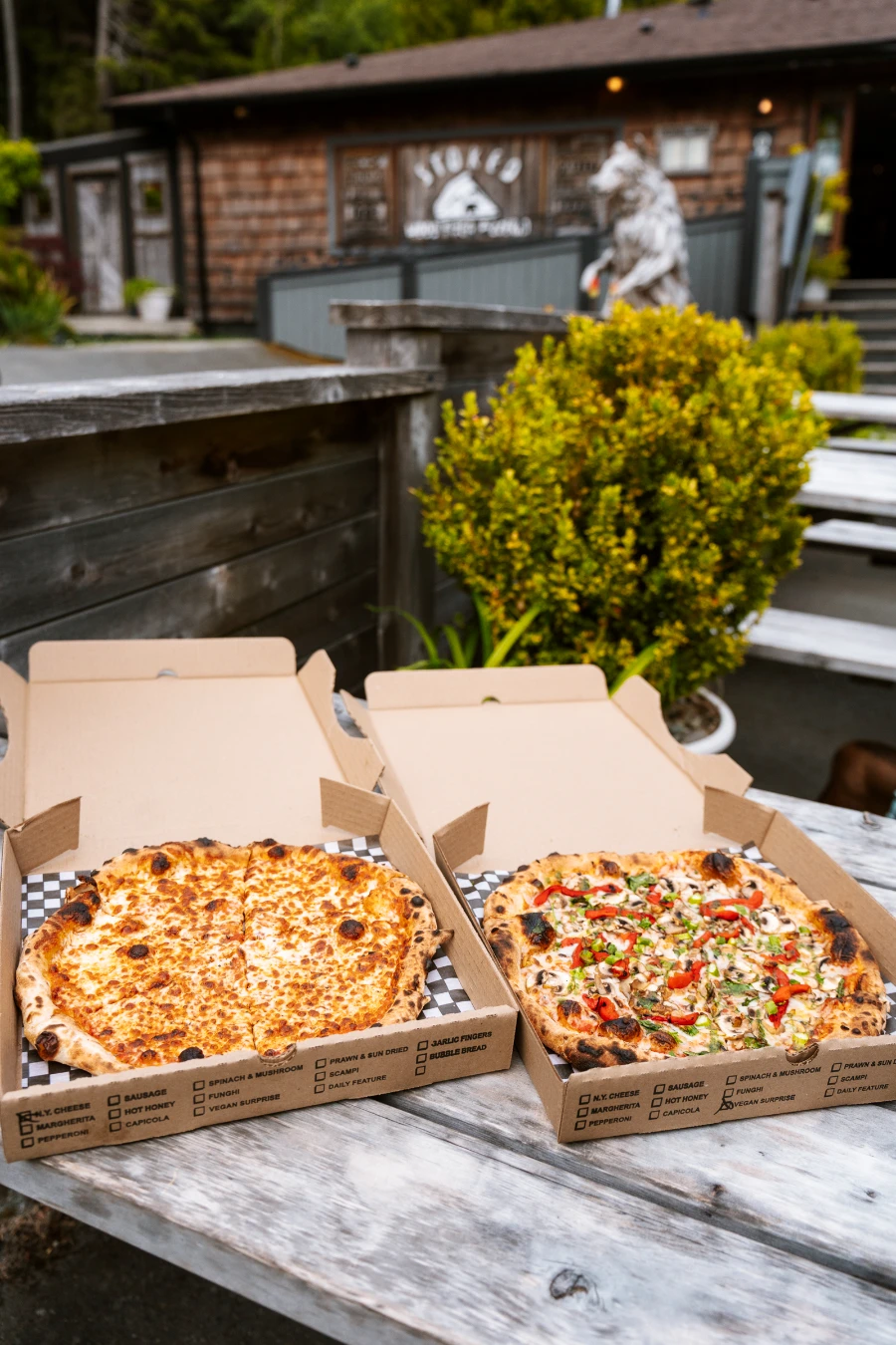 Two large pizzas in wooden take-away boxes on wooden picnic tables