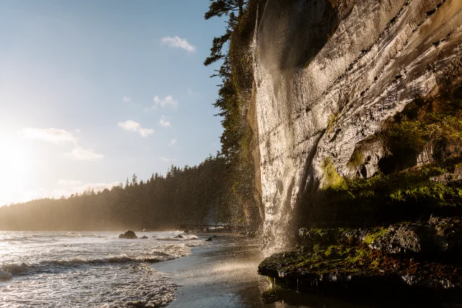 A misty waterfall pours over a cliff into the sea on a sandy beach below at Mystic Beach
