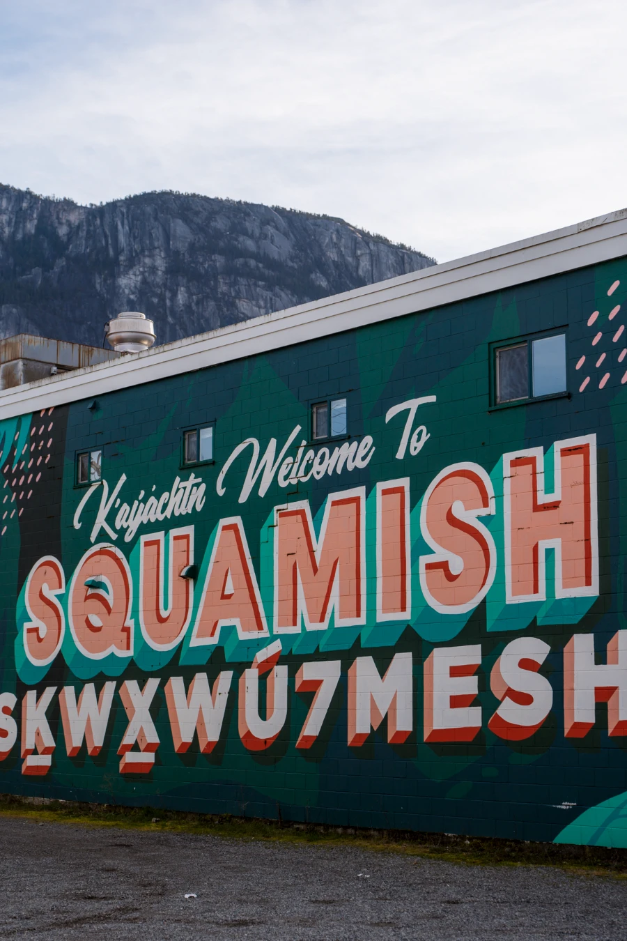 A mural on a building facing a parking lot says ‘Welcome To Squamish’ with the Stawamus Chief towering behind it