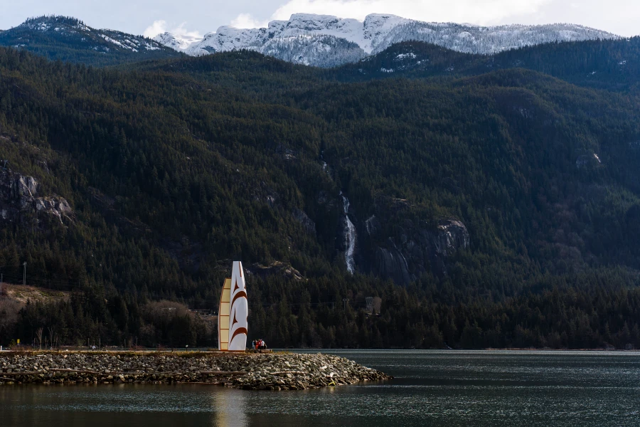 A rocky peninsula sticks out into the Howe Sound with a white monument built at the end of it, with Shannon Falls pouring over the side of the mountain across the Howe Sound