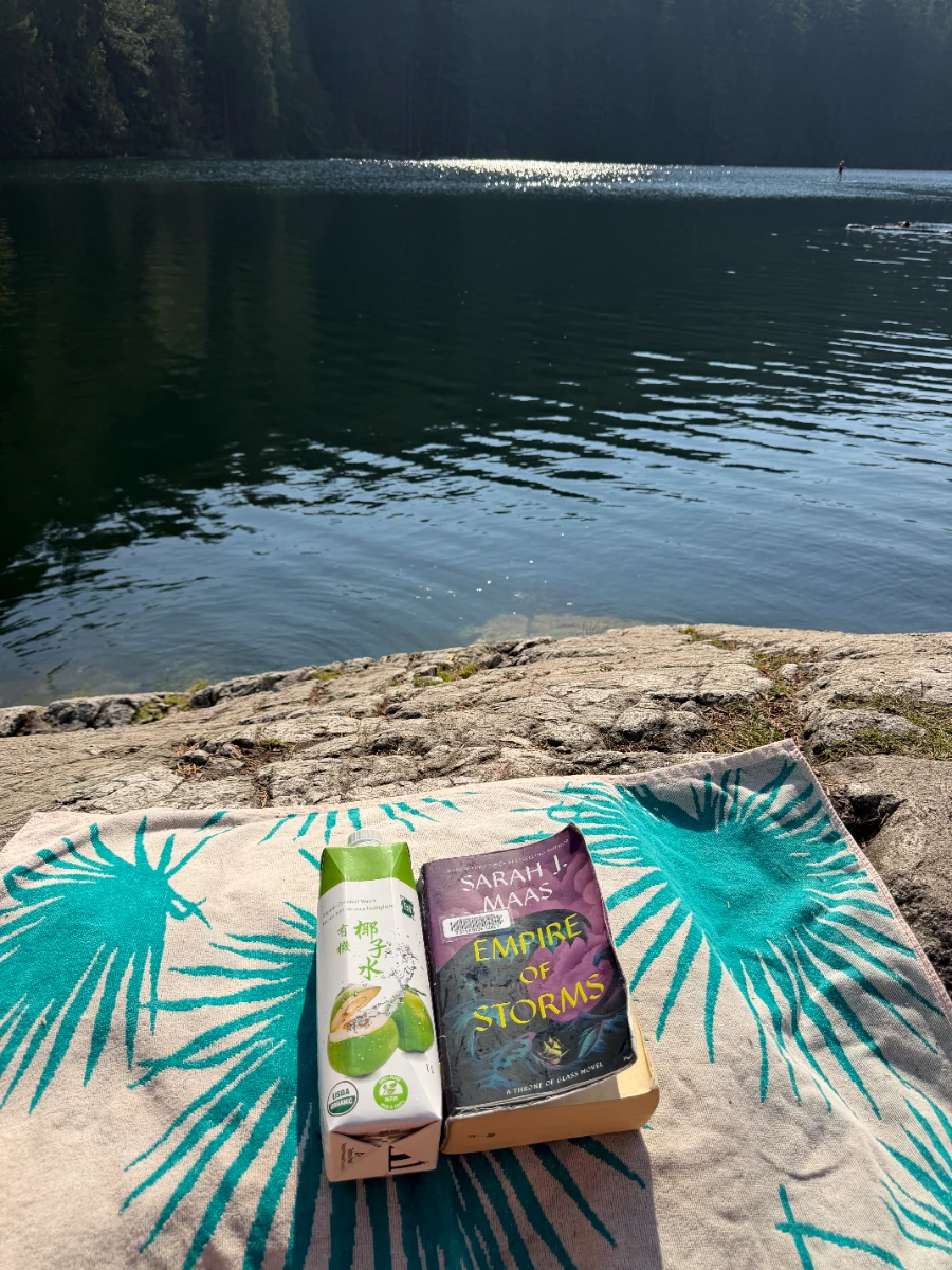 A book and a coconut water laid on a towel on a rock that is next to a small lake that is lined on all sides with trees
