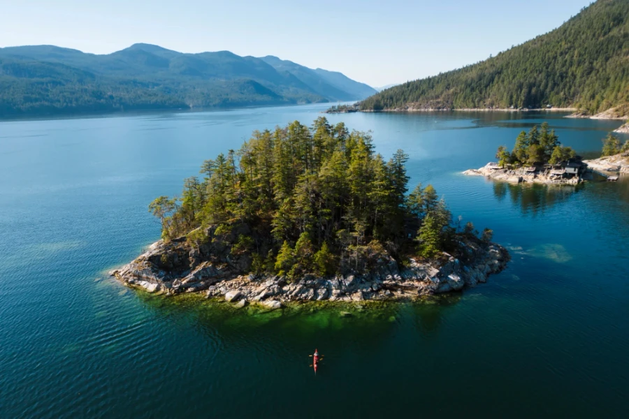 2 people in a red kayak pointed towards a small rocky island that is covered and trees in the Sechelt Inlet