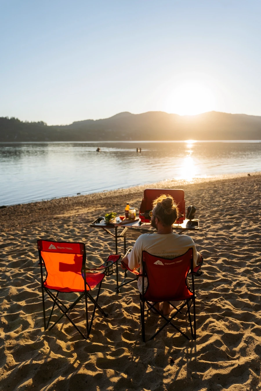 A man sits on a red camp chair on a sandy beach with a table in front of him with a camp stove on it