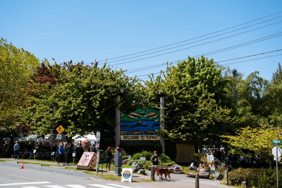 A sign reads welcome to Tofino next to a large crowd entering a park filled with tents at the Tofino Farmers Market