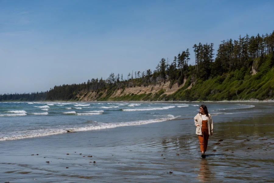 A woman walk along the wet sand of Florencia Bay in Tofino with Gentle waves rolling in to her left and massive sandstone cliffs behind her