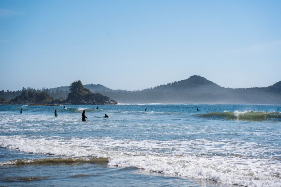 Mist rises above a collection of about a dozen surfers who are spread out across a large beach area with large waves crashing behind them on Long Beach in Tofino