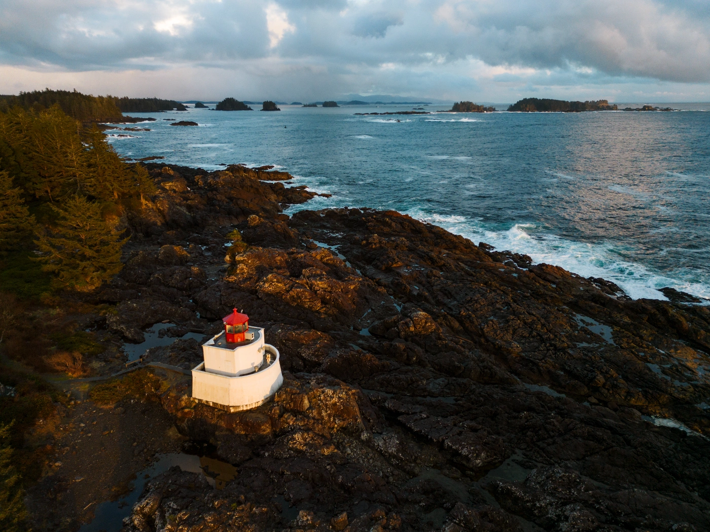 A red and white lighthouse glows in sunset light along a rocky tree lined coast