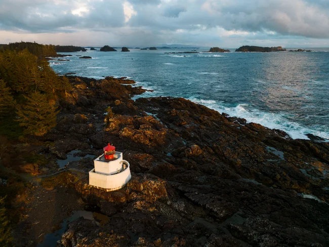 A red and white lighthouse glows in sunset light along a rocky tree lined coast