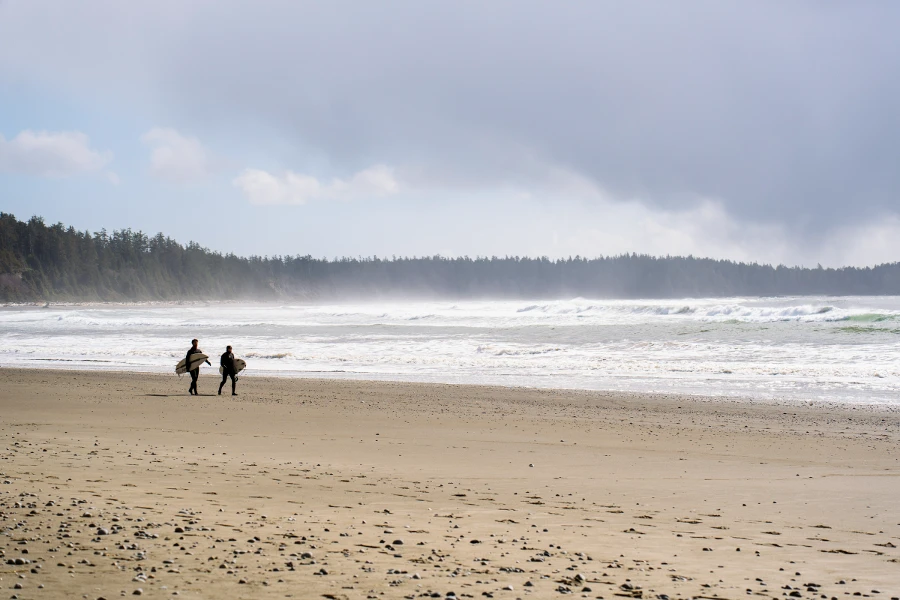 Two surfers walk with their boards under their arms along a wide white sand beach that is lined by misty rainforest near Ucluelet on Vancouver Island
