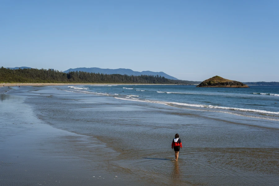 A woman stands in ankle deep water on an expansive beach as lines of waves roll in
