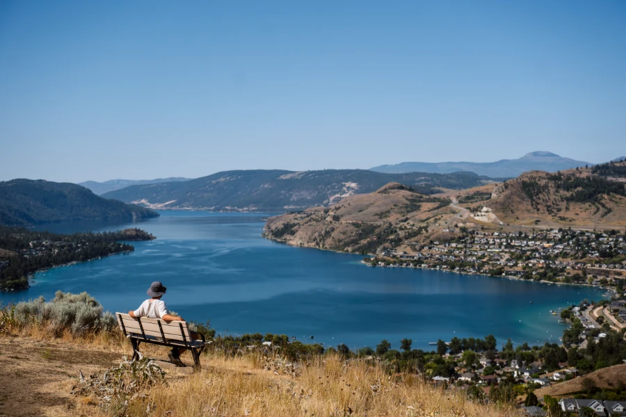A man in a sunhat sits on a wooden bench at the top of a huge hill covered in dry grass looking down at Kalamalka Lake