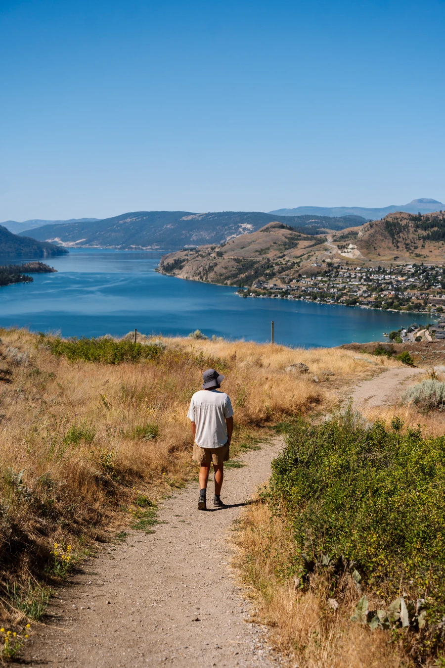 A man walks down a narrow dirt path surrounded on both sides by tall dry grass with the large blue mass of Kalamaka Lake in front of him